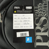 A close-up of a black PRS guitar body with a hangtag showing product details, model S2 Custom 24, and a blue S2 logo sticker. The tag is next to a silver and black PRS headstock section.