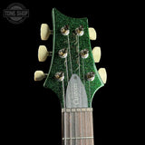 Close-up of a guitar headstock with a glittering dark green finish, cream-colored tuning pegs, and a silver maple neck against a black background.