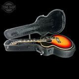 A 1930s-style acoustic guitar with a sunburst finish resting inside an open black hard case on a black background. The guitar features a warm orange-to-brown gradient body, a black neck, and decorative headstock detailing.