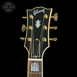 Close-up of a Gibson acoustic guitar headstock with gold tuning pegs, a black faceplate, and a dark wood fretboard on a black background.