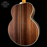 Close-up of the back of an acoustic guitar showing a rich dark brown wood grain finish, cream binding, and a portion of the neck at the top, set against a solid black background with a Tone Shop watermark in the upper left.