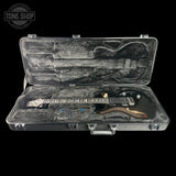 A black electric guitar with a metallic bronze headstock and gold hardware, displayed inside an open gray hard case with gray foam padding. The case is set against a solid black background, with a Tone Shop logo watermark in the top-left corner.