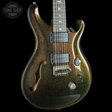 A close-up of a double-cutaway electric guitar with a dark metallic gold glitter finish, featuring black humbucker pickups and chrome hardware, set against a solid black background.