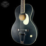 A black acoustic guitar with a glossy finish, white pickguard, and white binding, shown against a solid black background. A circular Tone Shop watermark is visible in the top-left corner.
