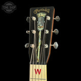 Close-up of the headstock of a vintage Martin & Co acoustic guitar against a solid black background. The headstock features a glossy black finish, a decorative vintage microphone inlay, six silver tuning pegs, and a light-colored neck with a red 'W' logo.
