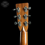 Close-up of the headstock of an acoustic guitar, showing six silver tuning pegs with ornate decorative engravings on a warm brown wood finish, set against a solid black background.