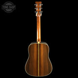 The back of a wooden acoustic guitar, showing a rich dark brown wood grain finish and a gold-toned center stripe, photographed against a solid black background with a Tone Shop watermark in the top-left corner.
