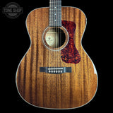 A close-up studio shot of an acoustic guitar body against a black background, showing a natural wood grain finish, a red tortoiseshell-style pickguard, and part of the neck. A Tone Shop watermark is visible in the top left corner.