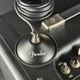 Close-up of a Fender amplifier footswitch with a coiled black cable, set against a black textured leather-like body with silver controls.