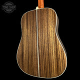Close-up of the back of an acoustic guitar showing a rich, dark brown wood grain finish with a light cream binding and center stripe, set against a solid black background.