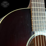 Close-up of an acoustic guitar body and neck showing a dark brown to black sunburst finish, gold-toned binding, and fingerboard on a black background. A Tone Shop watermark is visible in the top-left corner.