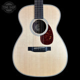 A close-up studio shot of an acoustic guitar body against a solid black background, featuring a natural flamed wood top, a dark reddish-brown pickguard, and a black bridge. A Tone Shop watermark is visible in the top left corner.