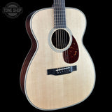 A close-up studio shot of an acoustic guitar body and upper neck against a solid black background. The guitar has a light natural wood top with a visible grain, a dark reddish-brown pickguard, a black bridge, and a dark wood side bout.
