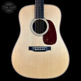 A front-facing acoustic guitar with a natural wood finish and a dark reddish-brown pickguard, shown against a solid black background with a Tone Shop watermark in the top-left corner.