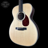 A Taylor acoustic guitar with a natural wood finish and a dark reddish-brown pickguard, shown against a solid black background with a Tone Shop watermark in the top left corner.
