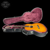 An acoustic guitar with a sunburst finish displayed inside an open black hard case with a dusty rose velvet lining, set against a solid black background.