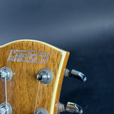 Close-up of the headstock of a Gretsch acoustic guitar, showing the wood grain, silver Gretsch logo, tuning pegs, and tuning keys against a dark blue background.