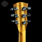 Close-up of the headstock of a guitar with a natural wood grain finish and six black tuning pegs, shown against a solid black background with a Tone Shop watermark in the top left corner.