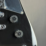 Close-up of a glossy black electric guitar body showing two black control knobs with white markings, a chrome pickguard, and a silver edge trim against a neutral gray background.