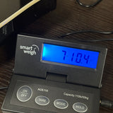 A close-up of a smart weigh ACE110 digital scale sitting on a dark wooden table. The scale is black plastic with a blue backlit LCD screen displaying '710.