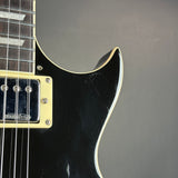 Close-up of a black electric guitar body showing the edge of the contoured headstock, a cream-colored binding, a gold-toned humbucker pickup, and part of the dark fretboard against a neutral gray background.