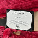 A Gibson Custom Certificate of Authenticity card inside a black folder, placed on plush red fabric with part of a wooden guitar body visible at the right edge.