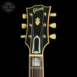 Close-up of a Gibson electric guitar headstock with gold tuning pegs, a white pickguard, and a dark wood fretboard on a black background.