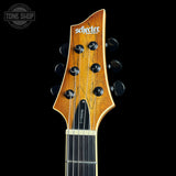 Close-up of the headstock of a Schecter electric guitar against a black background, showing the amber wood finish, six black tuning pegs, and the Schecter logo.