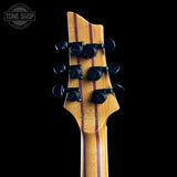 Close-up of the headstock of a guitar with a natural wood finish, featuring two dark red wood stripes running vertically and six black tuning pegs. The Tone Shop logo is visible in the top left corner.