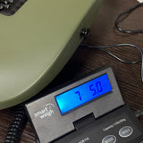 A close-up of a digital smart weigh scale displaying '7.50 kg' in blue, placed on a dark wood surface next to an olive green guitar body.