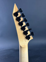 Close-up of the headstock of a guitar with a natural wood finish and black tuning pegs, set against a dark blue-gray background.
