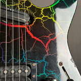 Close-up of a black electric guitar body featuring a vibrant, multicolor crackle pattern in red, orange, yellow, green, blue, and purple over a distressed black finish.