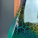 Close-up of a glossy black guitar body with a vibrant crackle rainbow pattern in blue, green, yellow, and purple, featuring a small clear bubble on the gloss finish.