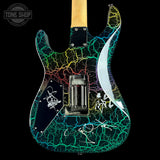 Custom electric guitar body featuring a vibrant multicolor crackle pattern in green, blue, yellow, and red over a black base, with white hand-painted signatures and a light wood neck visible at the top, set against a solid black background.