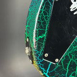 Close-up of the rear side of an electric guitar body featuring a black base with a vibrant green and blue crackle pattern, a black pickguard, and silver hardware against a gray background.