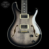 A close-up of a double-cutaway electric guitar against a solid black background. The guitar features a dark brown to black flamed maple top with a sunburst gradient, gold hardware, and white binding.