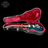 A blue electric guitar resting inside an open black hard case with red velvet lining, photographed against a solid black background. A Tone Shop watermark is visible in the top-left corner.