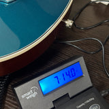 A close-up of a guitar body connected to a digital smart scale on a dark wooden surface. The scale displays a weight of 71.