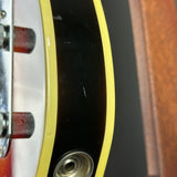Close-up detail of an electric guitar body showing a black floyd-deadboard area with a yellow edge, silver hardware, and part of a wood-toned headstock.
