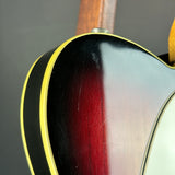 Close-up of the body of an electric guitar showing a sunburst finish that transitions from black to a deep red center, bordered by a gold edge and a white pickguard.