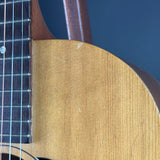 Close-up detail of an acoustic guitar showing the natural wood body, sound hole surround, neck, and dark fretboard against a dark blue background.