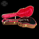 An acoustic guitar with a glossy black body and a dark brown neck, displayed inside an open brown hard case with a bright red velvet interior. The Tone Shop logo is visible in the top left corner.
