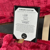 A Gibson Custom Certificate of Authenticity card inside a black frame, placed on a plush pink velvet surface with part of a tan strap and a wooden object visible.