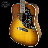 A close-up studio shot of an acoustic guitar body against a black background, showing a natural sunburst finish, a dark pickguard with blue and gold floral inlay, and part of the neck. A Tone Shop watermark is visible in the top-left corner.