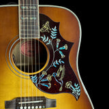 Close-up of a Yamaha acoustic guitar body featuring a natural wood finish, a dark tortoiseshell pickguard with blue and gold bird and floral inlay, and part of the fretboard.