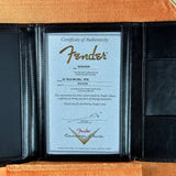 A Fender-branded Certificate of Authenticity document displayed inside an open black leather folder on an orange textured background.