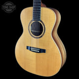 A natural wood acoustic guitar shown from a three-quarters angle against a solid black background, with a Tone Shop watermark in the upper left corner.