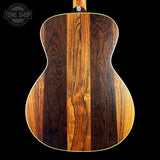 Close-up of the back of an acoustic guitar showing three vertical wood grain panels in dark brown, medium brown, and warm honey tones, with a white edge trim against a black background. A Tone Shop watermark is visible in the top left corner.
