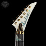 Close-up of a white electric guitar headstock with gold tuning pegs and a blue-green iridescent script logo reading 'Jackson' on a black background. A Tone Shop watermark is visible in the top-left corner.