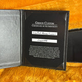 A black Gibson Custom Certificate of Authenticity booklet shown open on a textured orange-brown furry surface. The right page contains printed white text and two handwritten white label areas, while the left page is a gray textured interior.